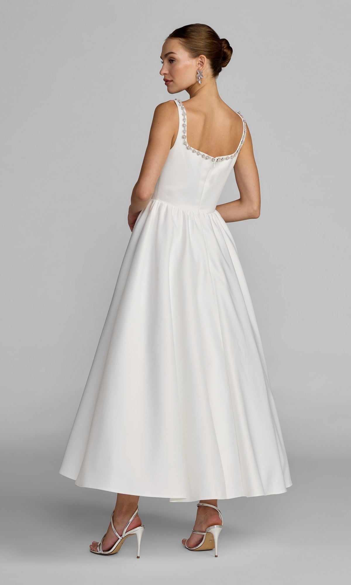 A woman stands sideways, glancing over her shoulder, wearing the Mac Duggal 56417 tea-length party dress—a sleeveless white satin design with a flared skirt and beaded neckline—paired with strappy heels and drop earrings.