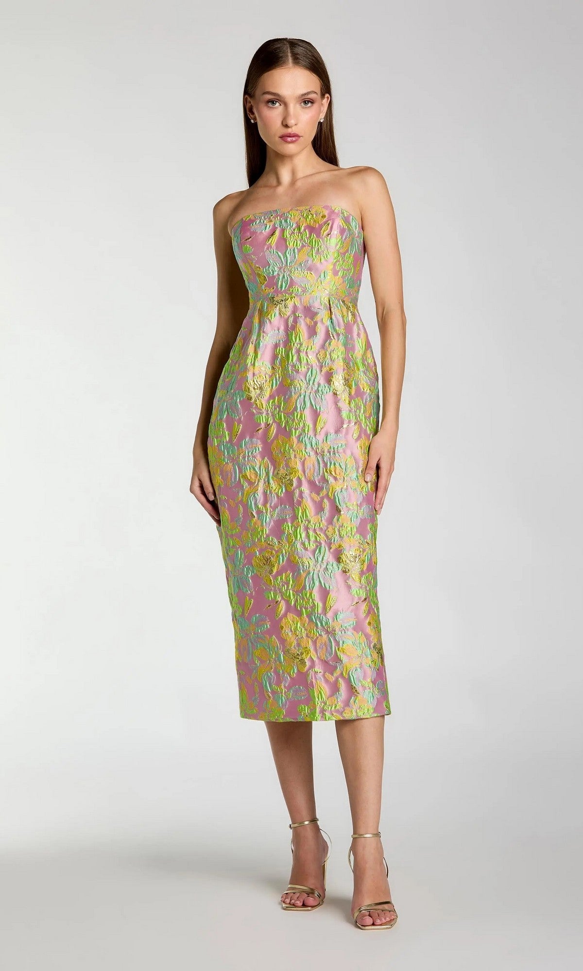 A woman wearing the Midi Wedding Guest Dress: Mac Duggal 56423, a strapless brocade creation in knee-length pastel pink adorned with green and yellow floral patterns, stands against a plain light gray background.