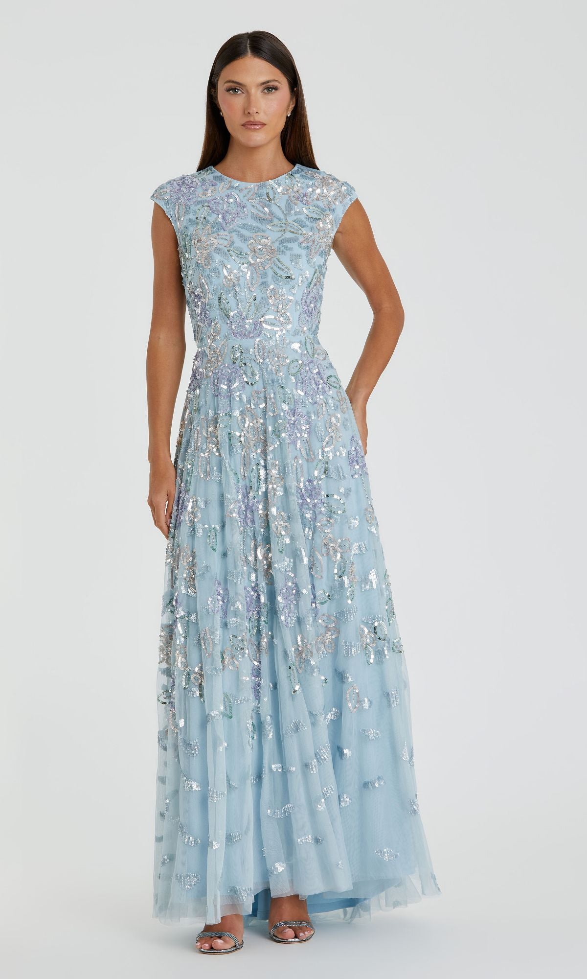 A woman wears the Mac Duggal 5647, a long blue sequin formal dress with cap sleeves, paired with silver open-toe heels against a plain background.