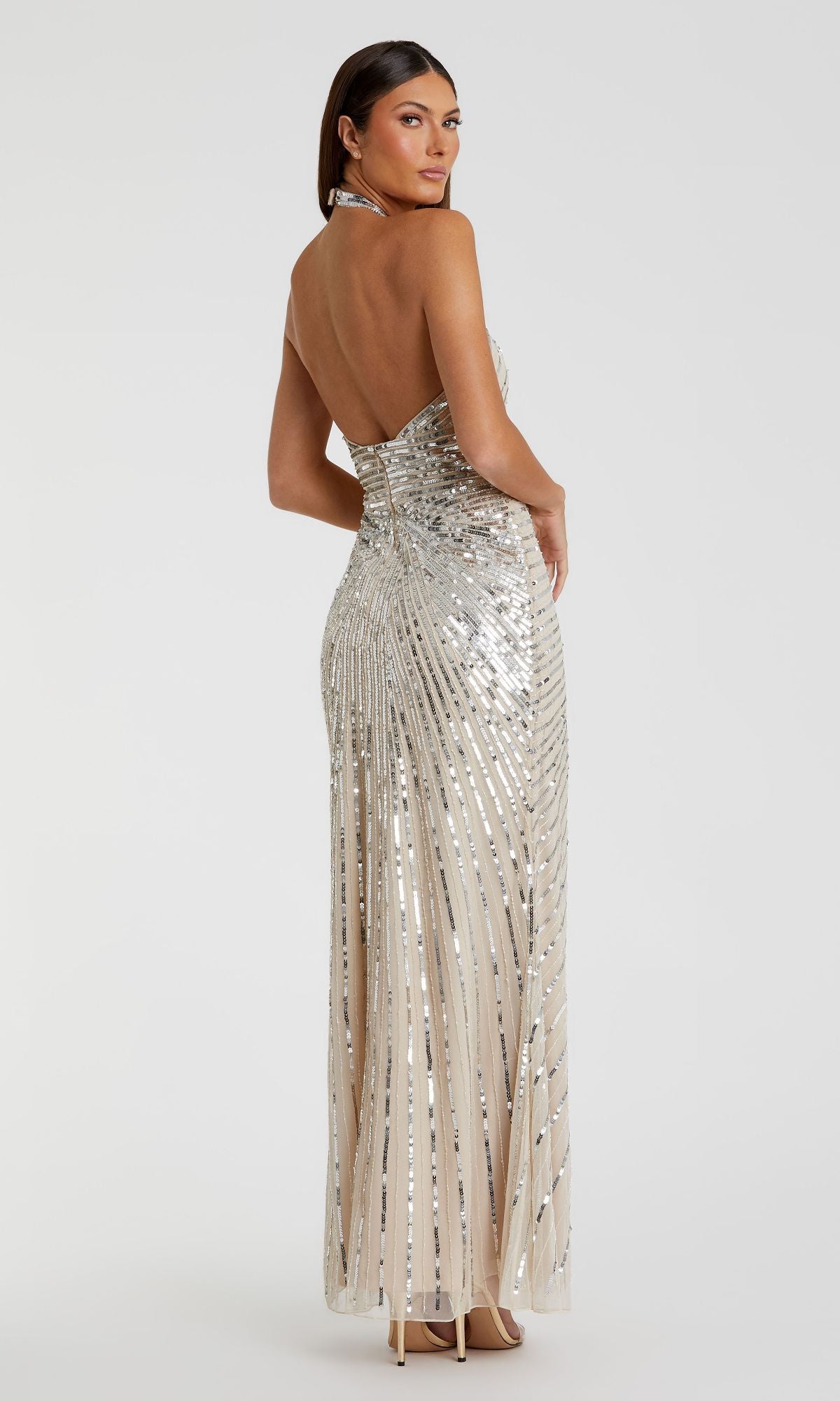 A woman poses in the Mac Duggal 5650, a floor-length silver sequin halter formal dress with an open back and nude heels, set against a plain white background.