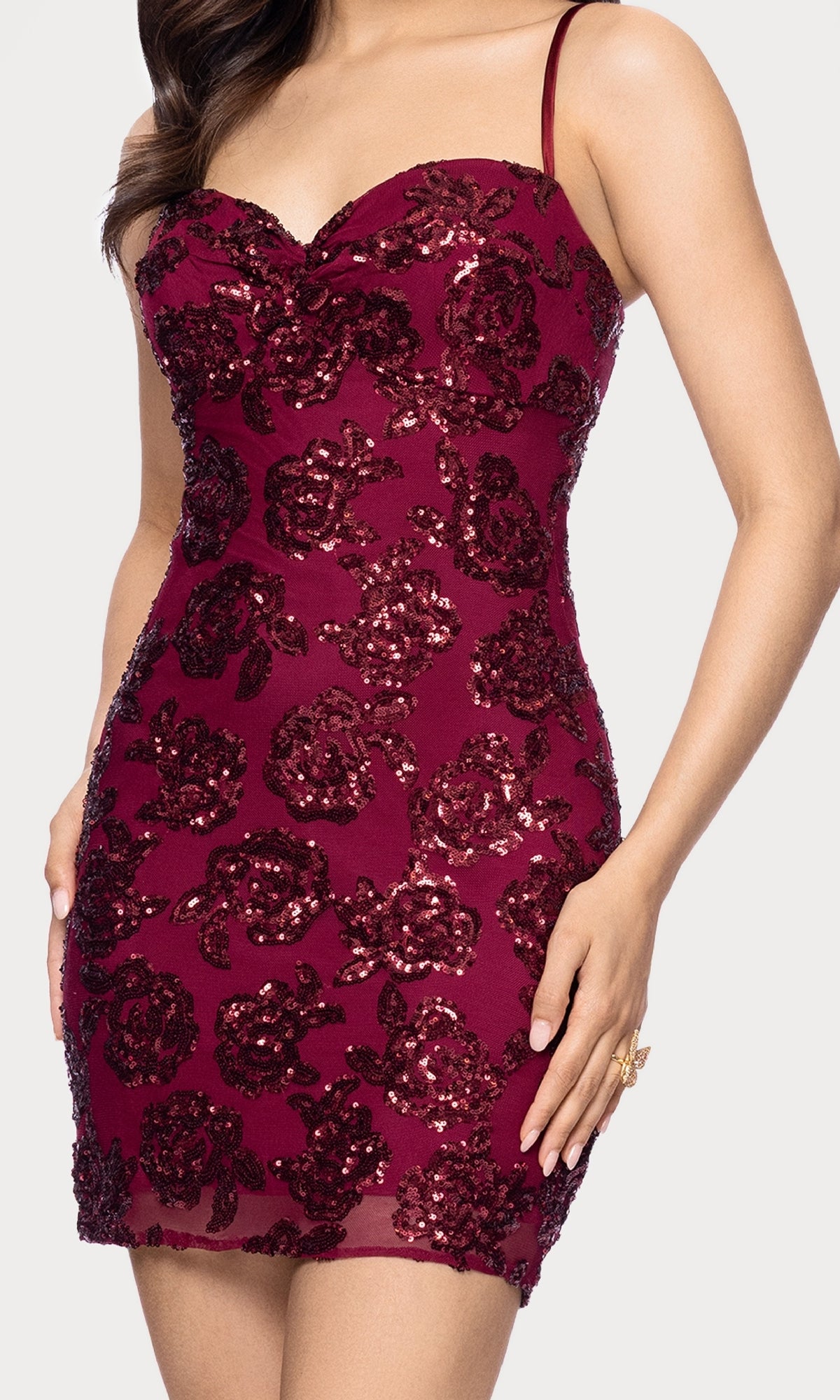 A woman poses with one hand on her hip wearing the Short Bodycon Hoco Dress: Blondie Nites 5679BN, a sleeveless, fitted burgundy mini dress featuring sequin floral patterns against a plain background.