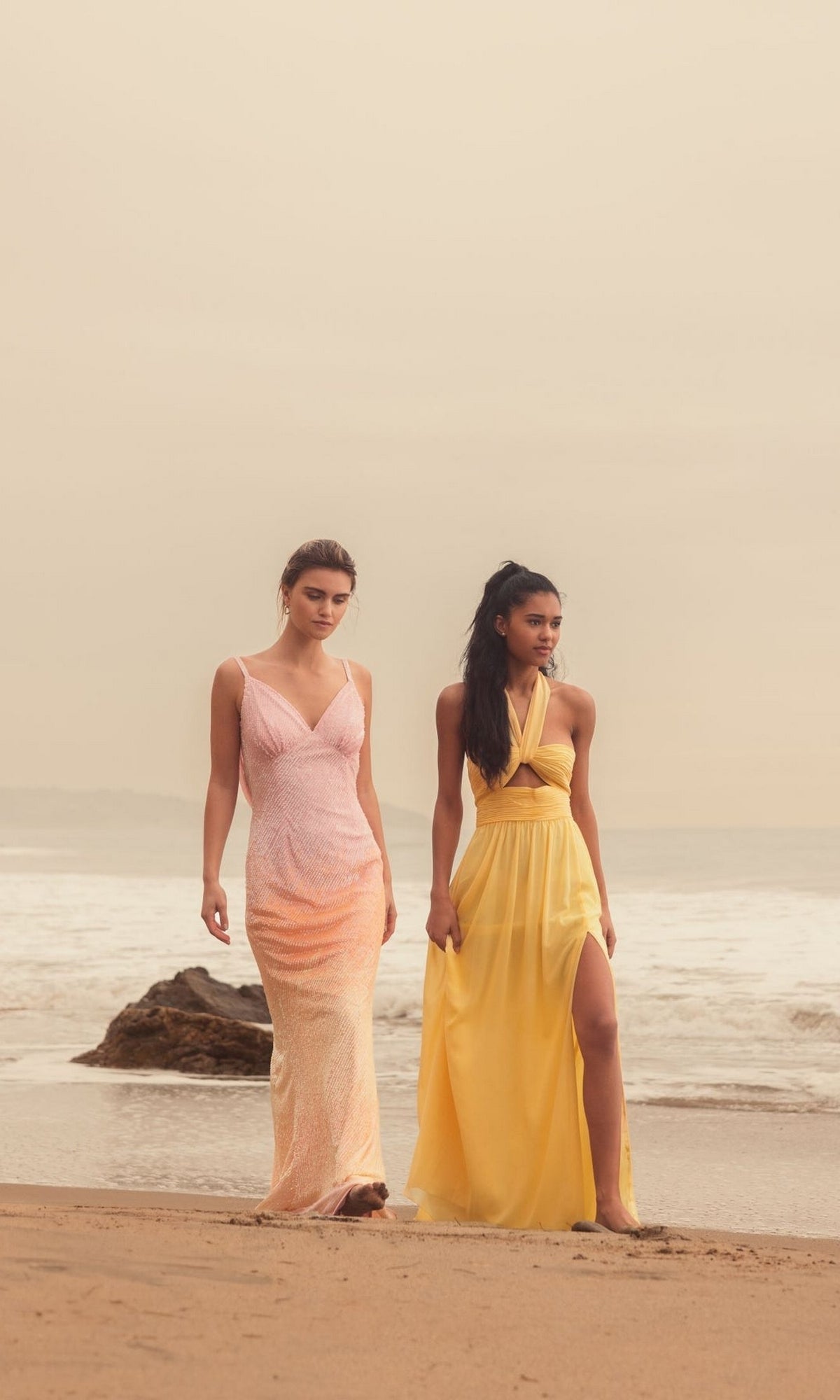Two women walk along a sandy beach under an overcast sky; one wears the Long Formal Dress 55898 by Mac Duggal, while the other wears a yellow ruched halter neck dress, with ocean and rocks visible in the background.