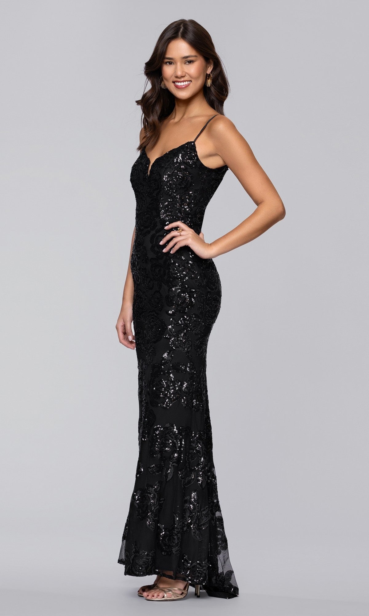 A woman poses confidently in a studio, wearing the Blondie Nites 5905BN long black prom dress—a floor-length, form-fitting gown with thin straps and floral sequin patterns.