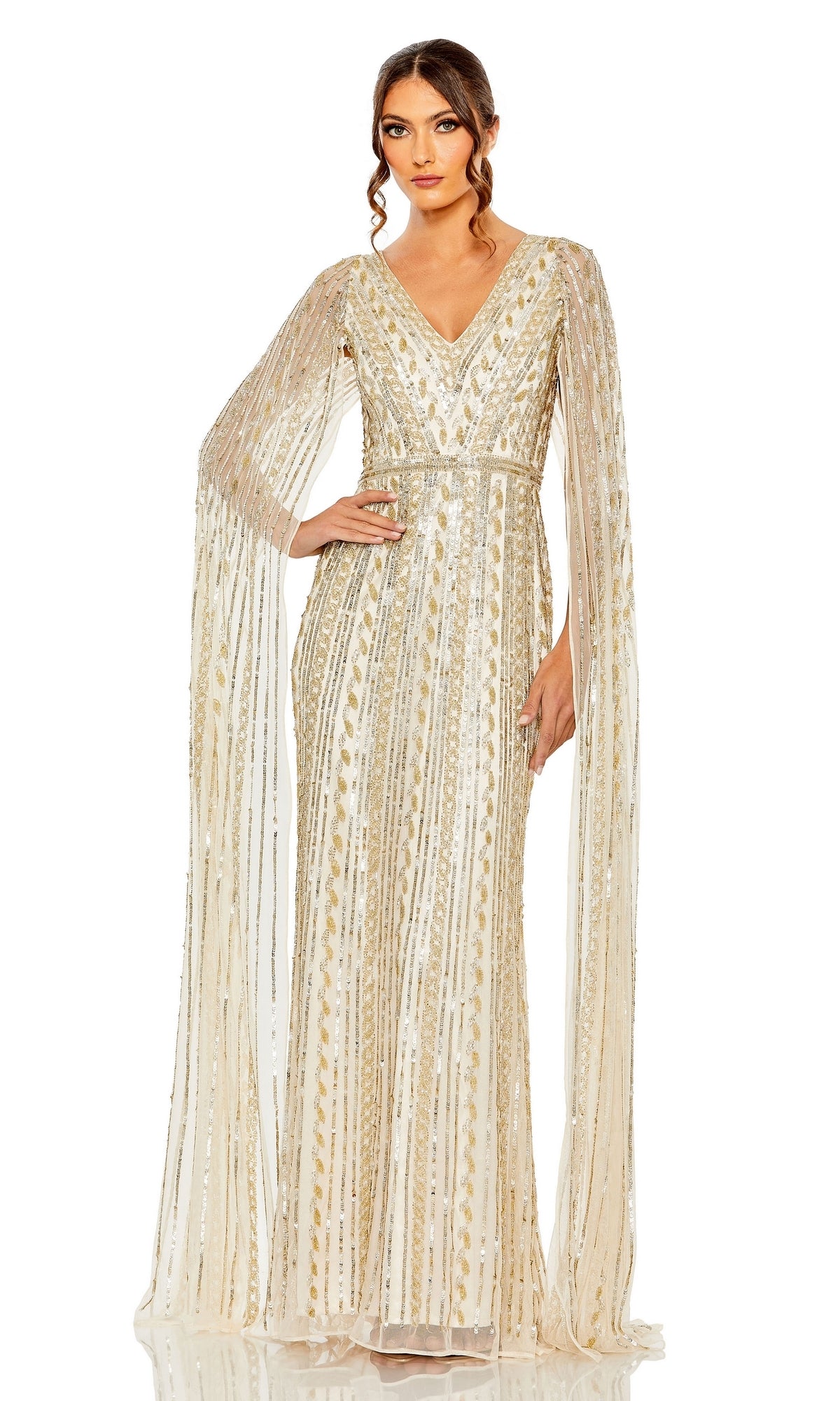 A woman poses with one hand on her hip, wearing the Long Formal Dress 5915 by Mac Duggal—a gold and white sequin gown featuring sheer, floor-length sleeves and a v-neckline—against a plain white background.