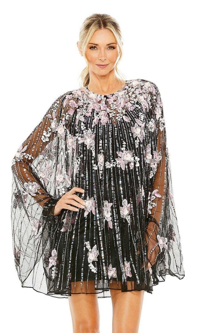 A woman wearing the Mac Duggal 5917 Beaded-Cape Short Hoco Dress, featuring a black short design with a sheer cape overlay embellished with pink and white floral embroidery, stands against a white background.