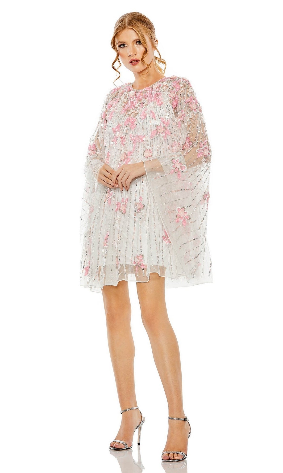 Woman wearing the Beaded-Cape Short Hoco Dress by Mac Duggal 5917— a sheer white dress with pink floral embroidery—paired with silver high-heeled sandals, standing against a plain white background.
