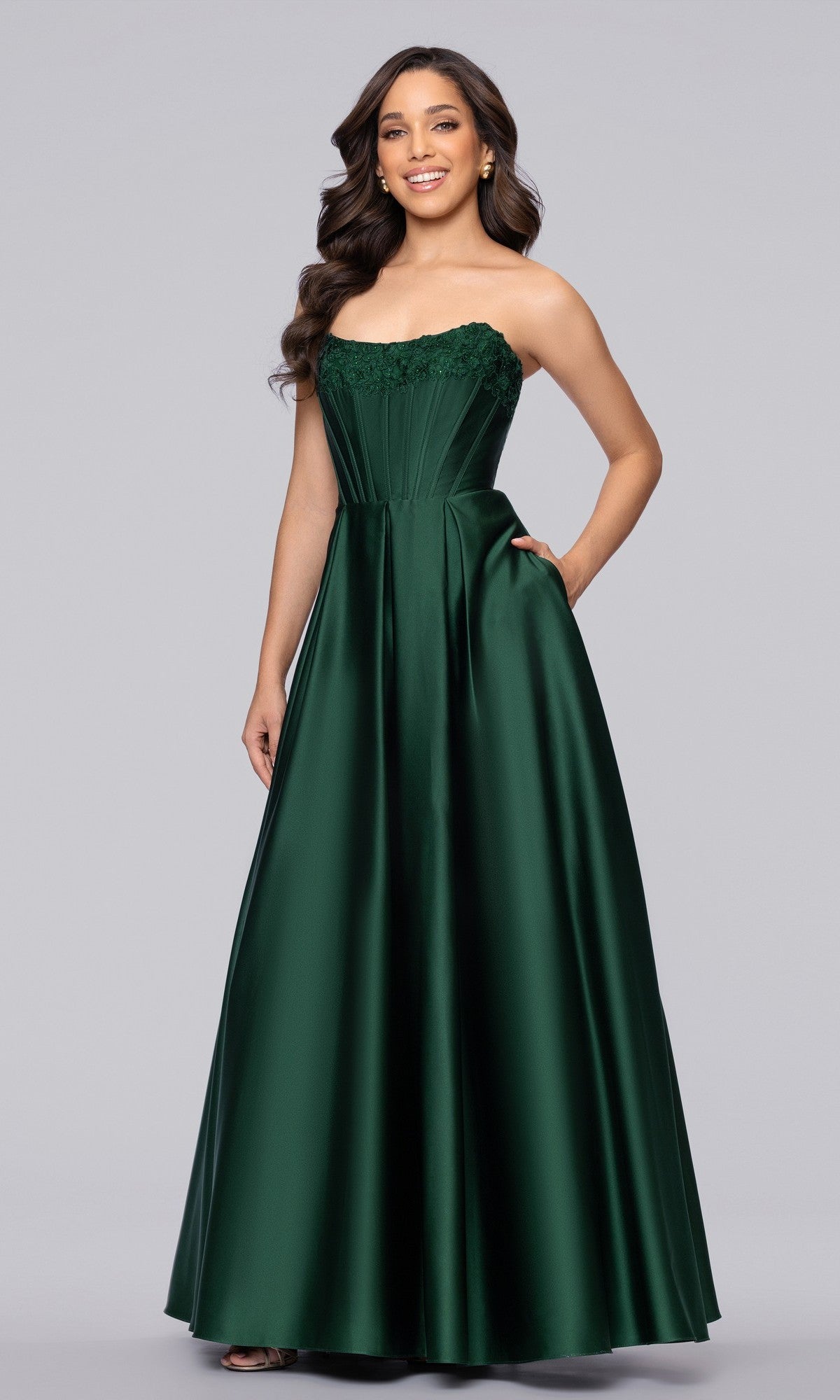 A woman stands against a plain light gray background wearing the Blondie Nites 5918BN long prom dress, featuring a strapless satin corset bodice and an emerald green floor-length skirt.