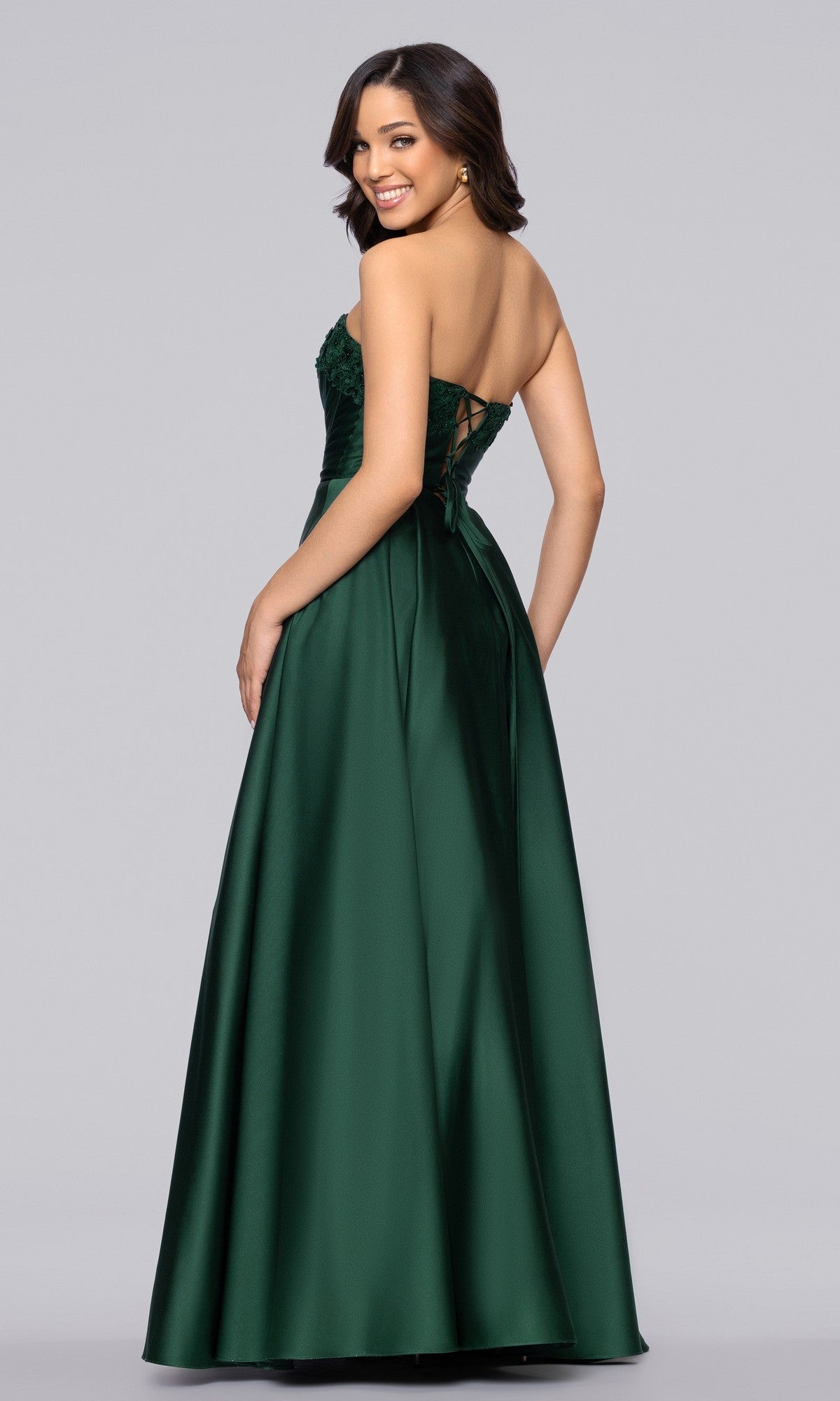 A woman stands smiling over her shoulder in the Blondie Nites 5918BN long prom dress, featuring a strapless satin design with a fitted corset bodice and lace-up back, against a plain background.