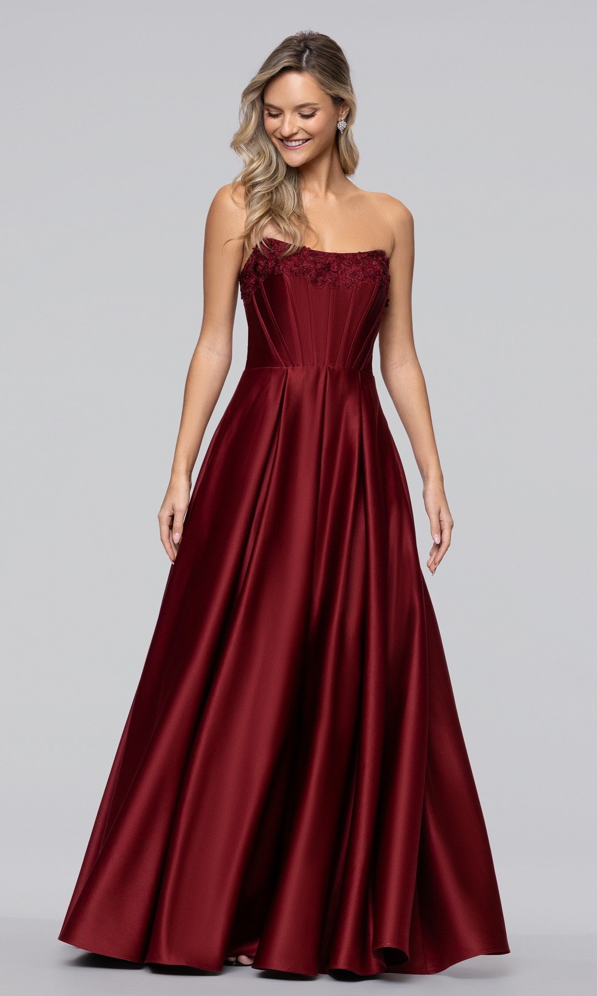 A woman stands against a plain gray background wearing the Blondie Nites 5918BN long prom dress, a burgundy satin strapless gown with a corset bodice, flared floor-length skirt, and subtle floral embroidery for an elegant finish.