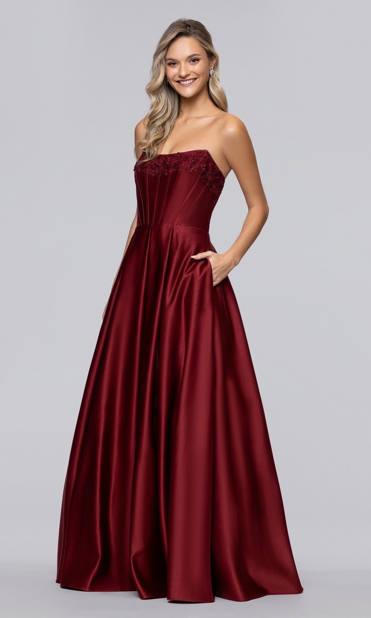 A woman stands against a light gray background wearing the Blondie Nites 5918BN long prom dress in wine—a strapless satin, floor-length gown with a fitted corset bodice and pockets.