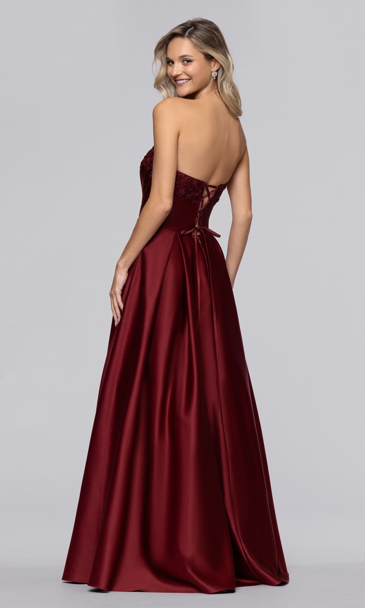 Smiling over her shoulder, a woman wears the Blondie Nites 5918BN long prom dress—a strapless, floor-length burgundy satin gown with a corset bodice and lace-up back detail.