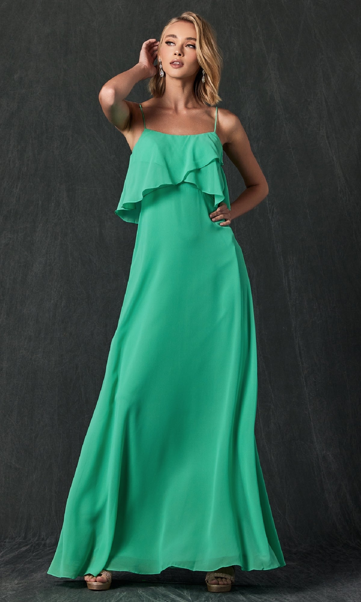 A woman in the Juliet 591 long, sleeveless green formal wedding-guest dress with spaghetti straps and a ruffled bodice stands against a dark textured background.