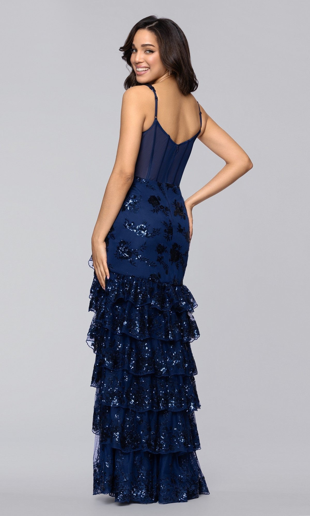 A woman in the Blondie Nites 5962BN long prom dress, featuring navy blue sequins and a tiered ruffle skirt, stands slightly turned to the side and smiling against a plain light gray background.