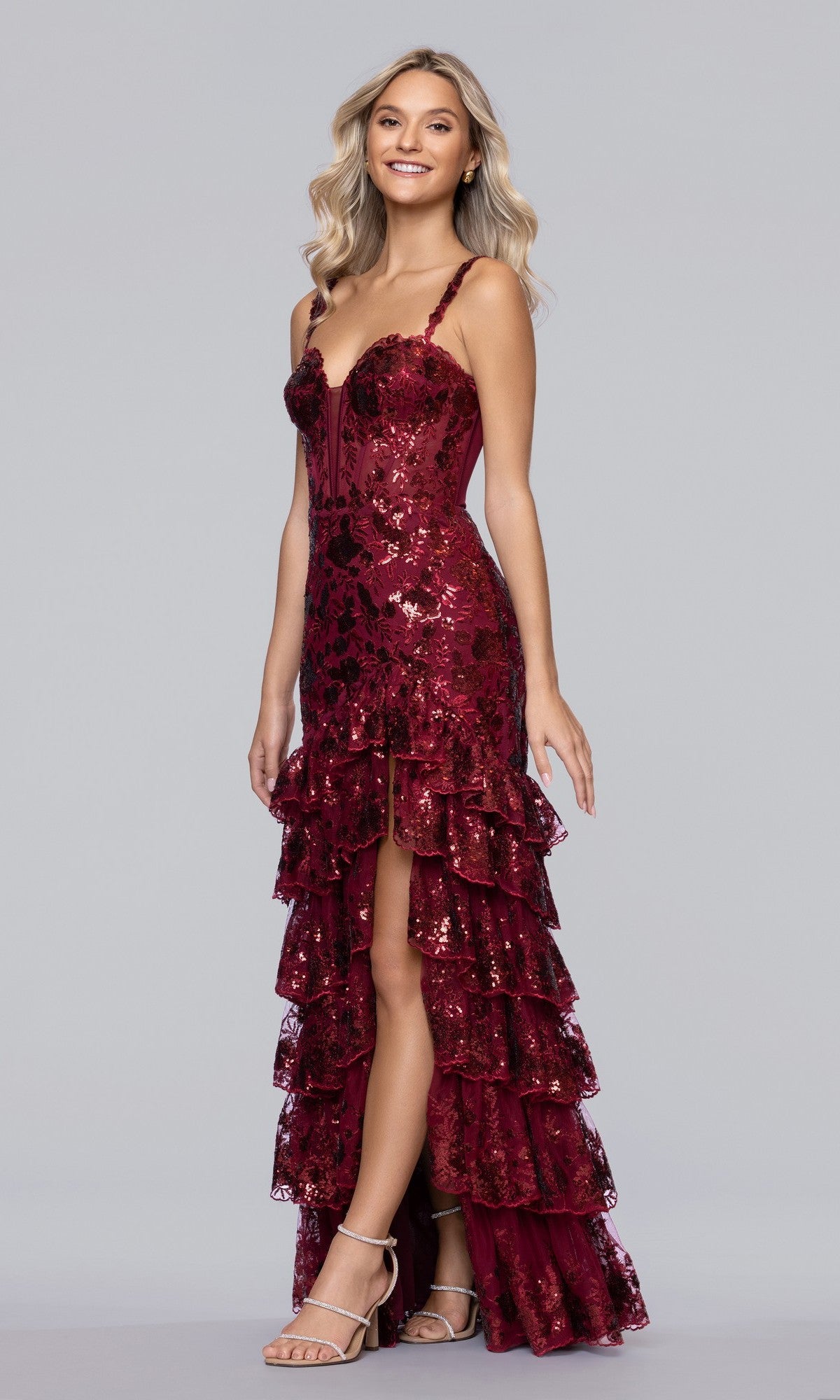 A woman in the Blondie Nites 5962BN, a sleeveless burgundy sequin prom dress with a tiered ruffle skirt and high slit, stands smiling against a plain gray background.