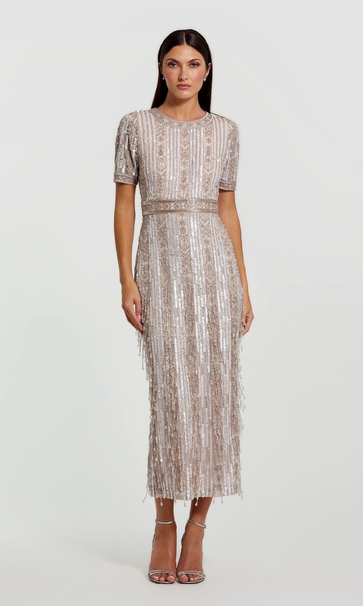 A woman models the Mac Duggal 5980 Midi Fringe Wedding-Guest Dress, featuring short sleeves, silver sequins, and fringe, paired with silver high-heeled sandals—an eye-catching semi-formal style against a plain backdrop.