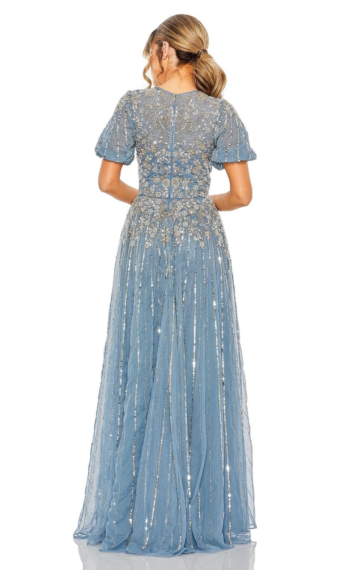 A woman with a ponytail is seen from the back wearing the Mac Duggal 5991 short sleeve beaded formal dress, a floor-length blue gown featuring silver embroidery and beadwork.