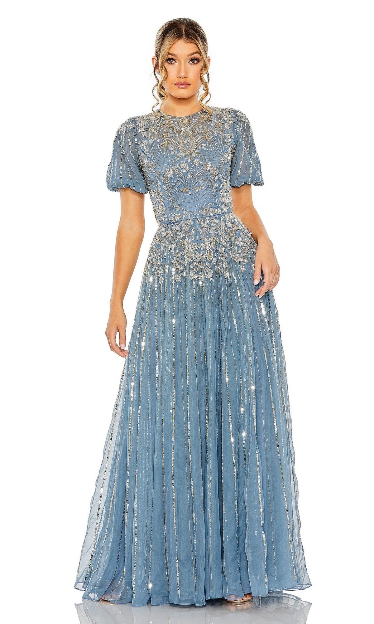 A woman wears the Mac Duggal 5991 Short Sleeve Beaded Formal Dress in blue, featuring intricate silver embroidery and a sheer overlay, standing against a white background.