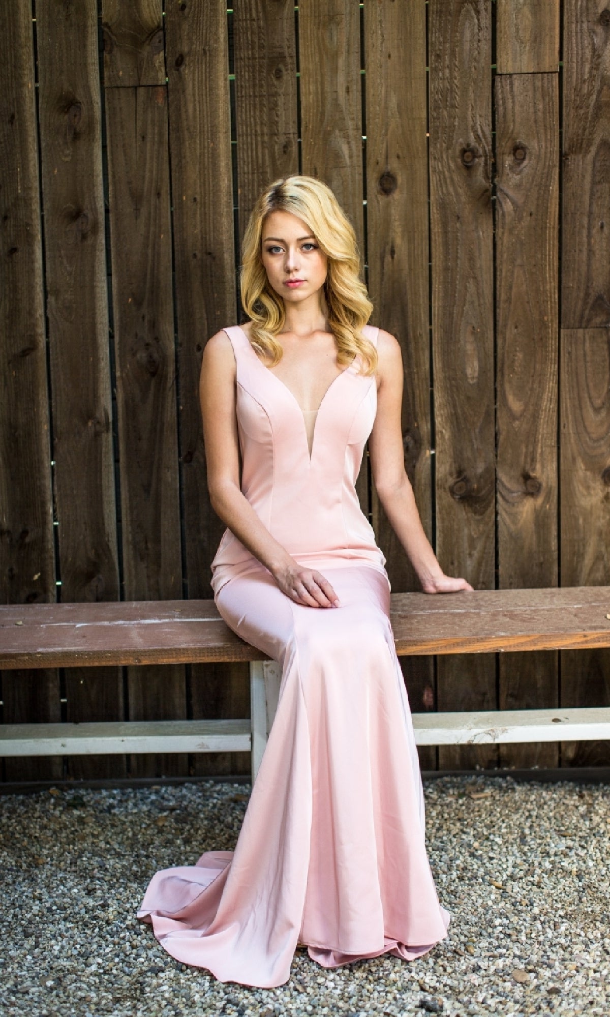 A woman wearing the Classic Long V-Neck Satin Prom Dress: Eureka 6010 sits on a wooden bench before a tall wooden fence, gazing slightly to the side.