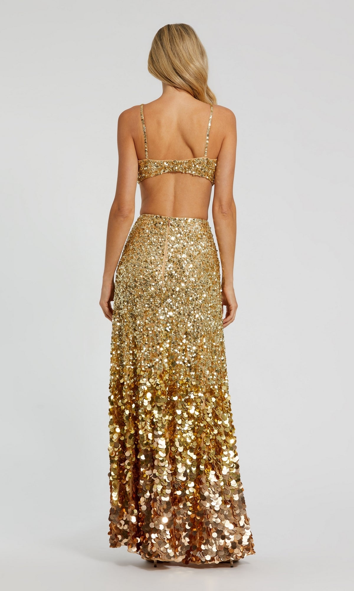 A woman stands facing away, wearing the Long Formal Dress 6077 by Mac Duggal—a gold sequin gown with an open back and a high-waisted, floor-length skirt.