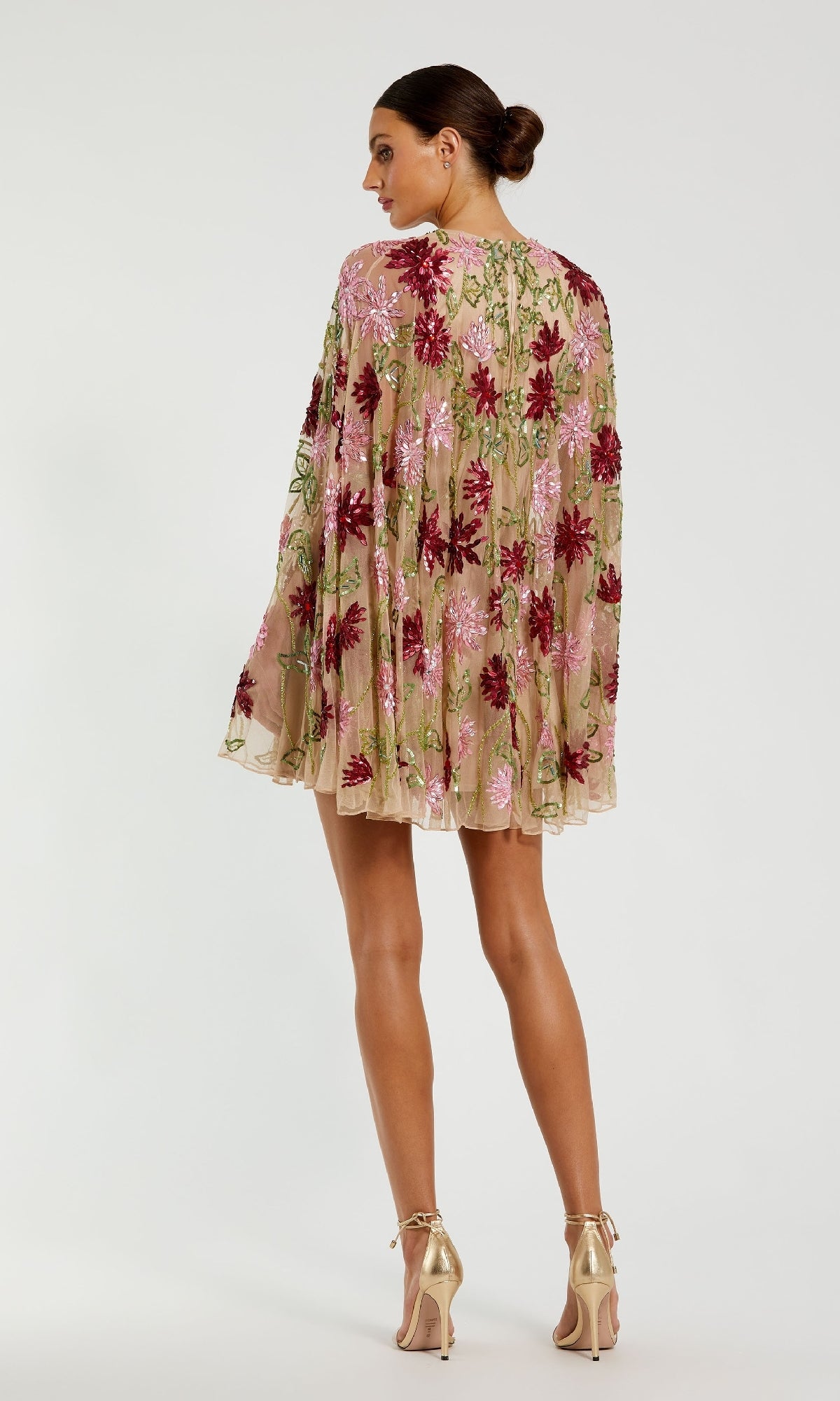 A woman stands facing away, wearing the Mac Duggal 6123 Sequin Short Wedding Guest Dress with long sleeves, pink and red floral embroidery on sheer beige fabric, styled with high heels.