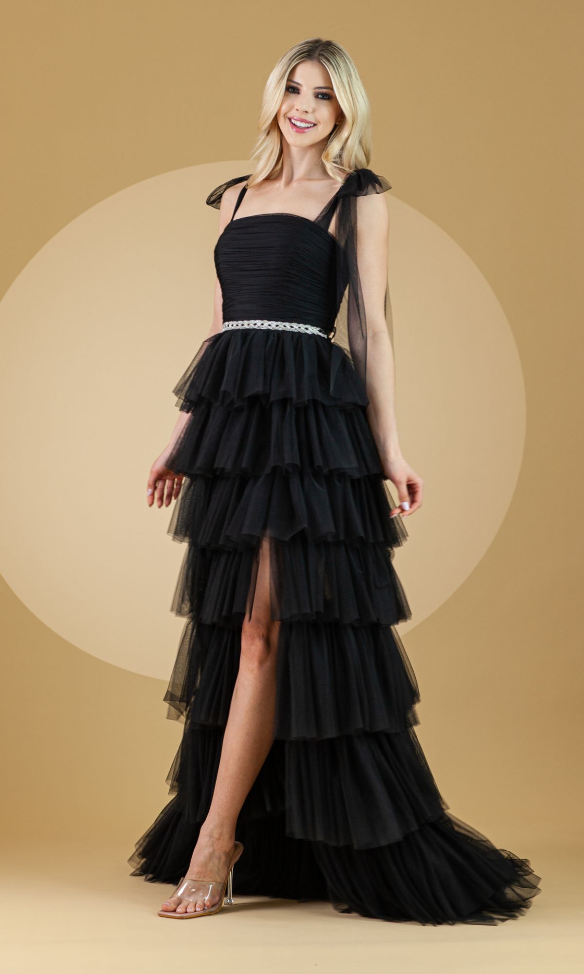 A woman in the Bow-Strap Ruffled Long Prom Ball Gown 6125, featuring tiered ruffles, a silver belt, and sheer bow shoulder straps, stands against a beige background, smiling.