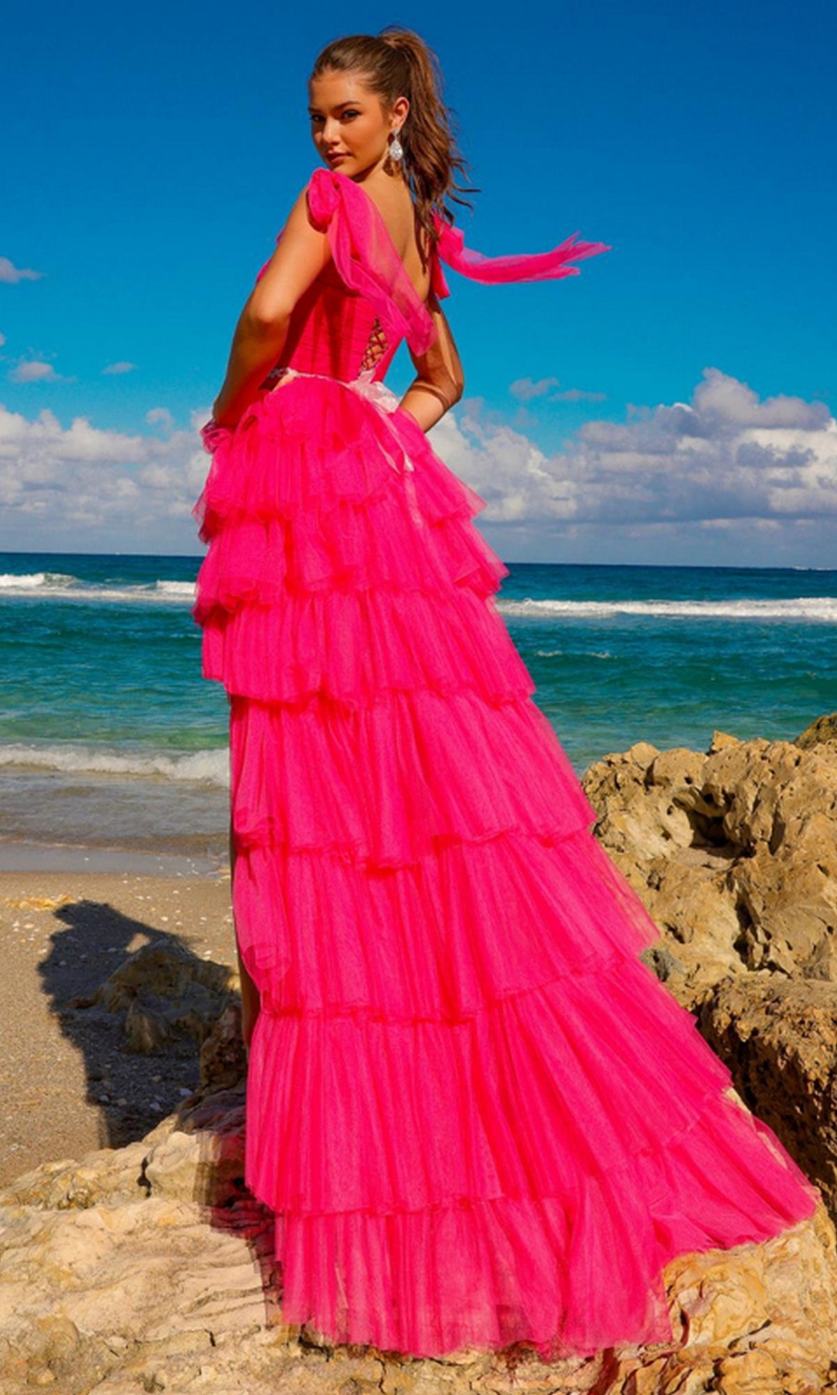 A woman stands on a rocky beach with the ocean and blue sky behind her, wearing the Bow-Strap Ruffled Long Prom Ball Gown 6125 in bright pink, featuring layered ruffles.
