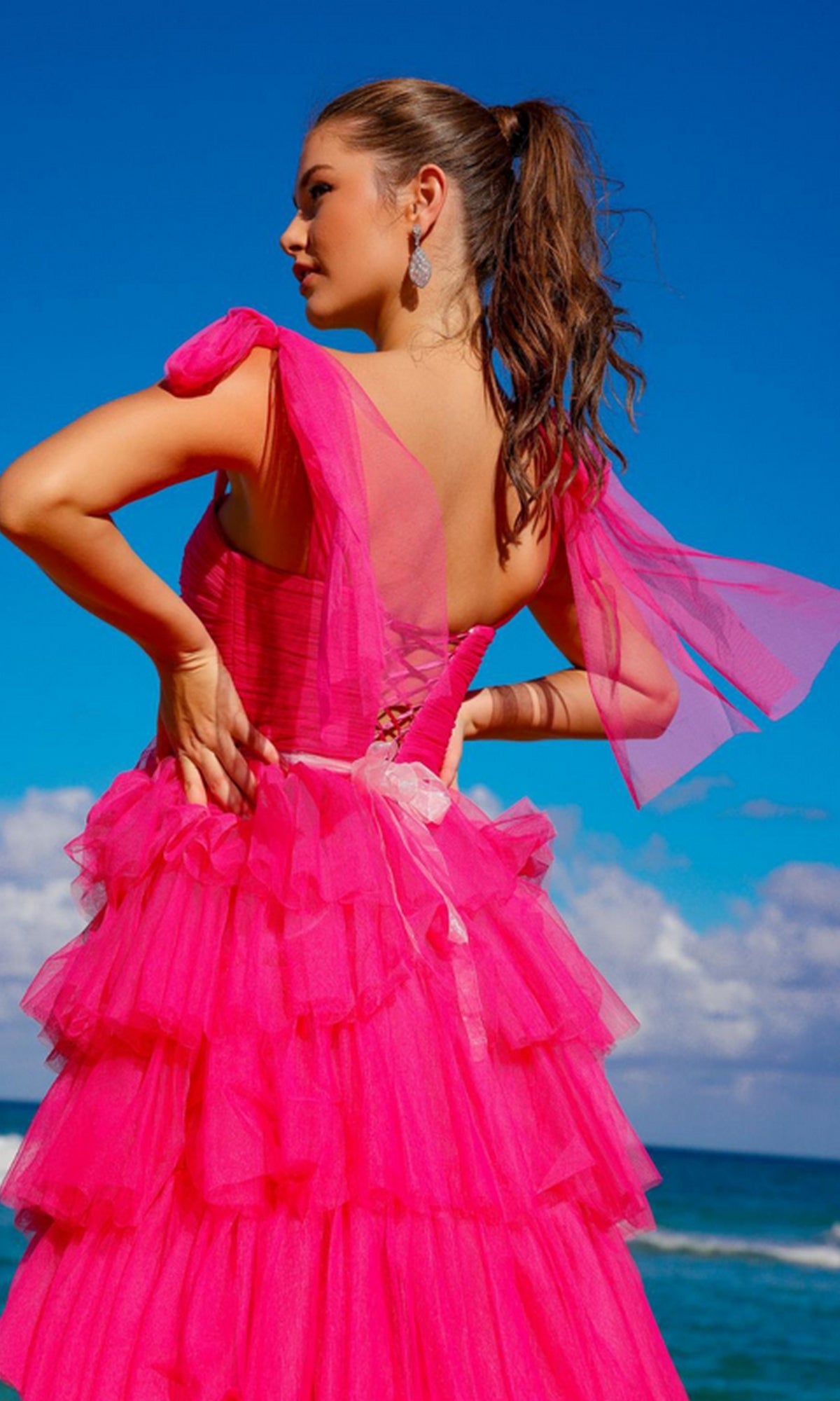 A woman in the Bow-Strap Ruffled Long Prom Ball Gown 6125, featuring sheer sleeves and bright pink ruffles, stands near the ocean under a blue sky, gazing to the side.