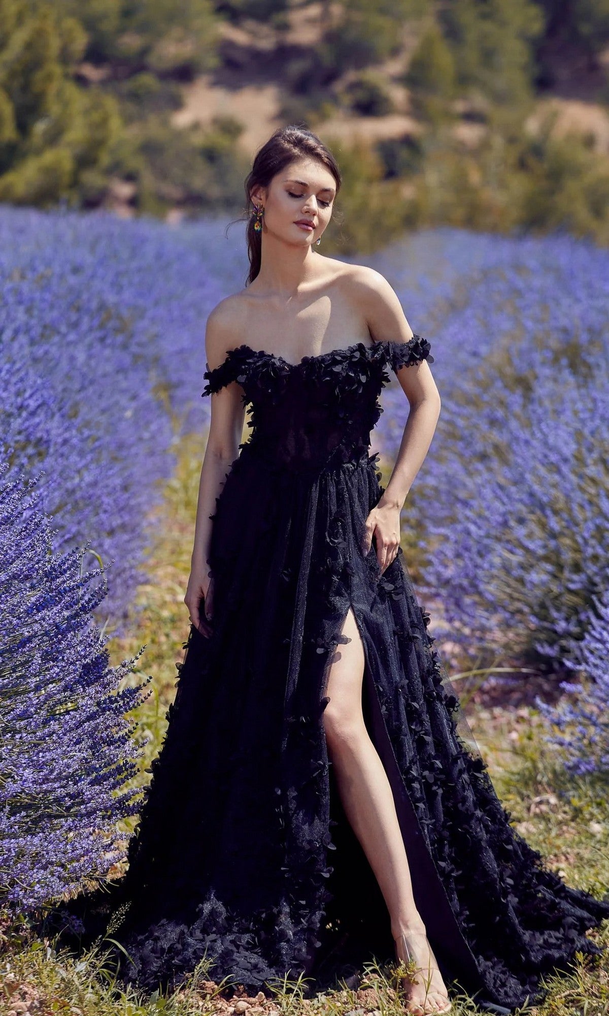 A woman wearing the Alyce 61308 black off-shoulder long floral dress with a thigh-high slit stands gracefully in a field of blooming lavender flowers.