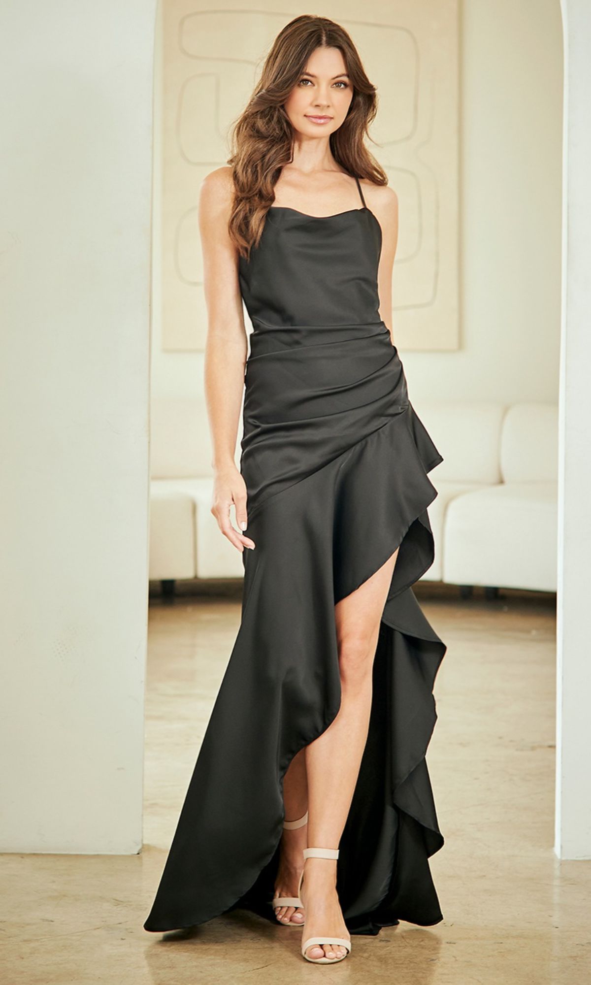 Indoors, a woman wears the Amelia Couture 6131 Strappy-Back Long Prom Dress with a black satin finish, featuring a high-low ruffled hem and an open back, styled with nude heels.