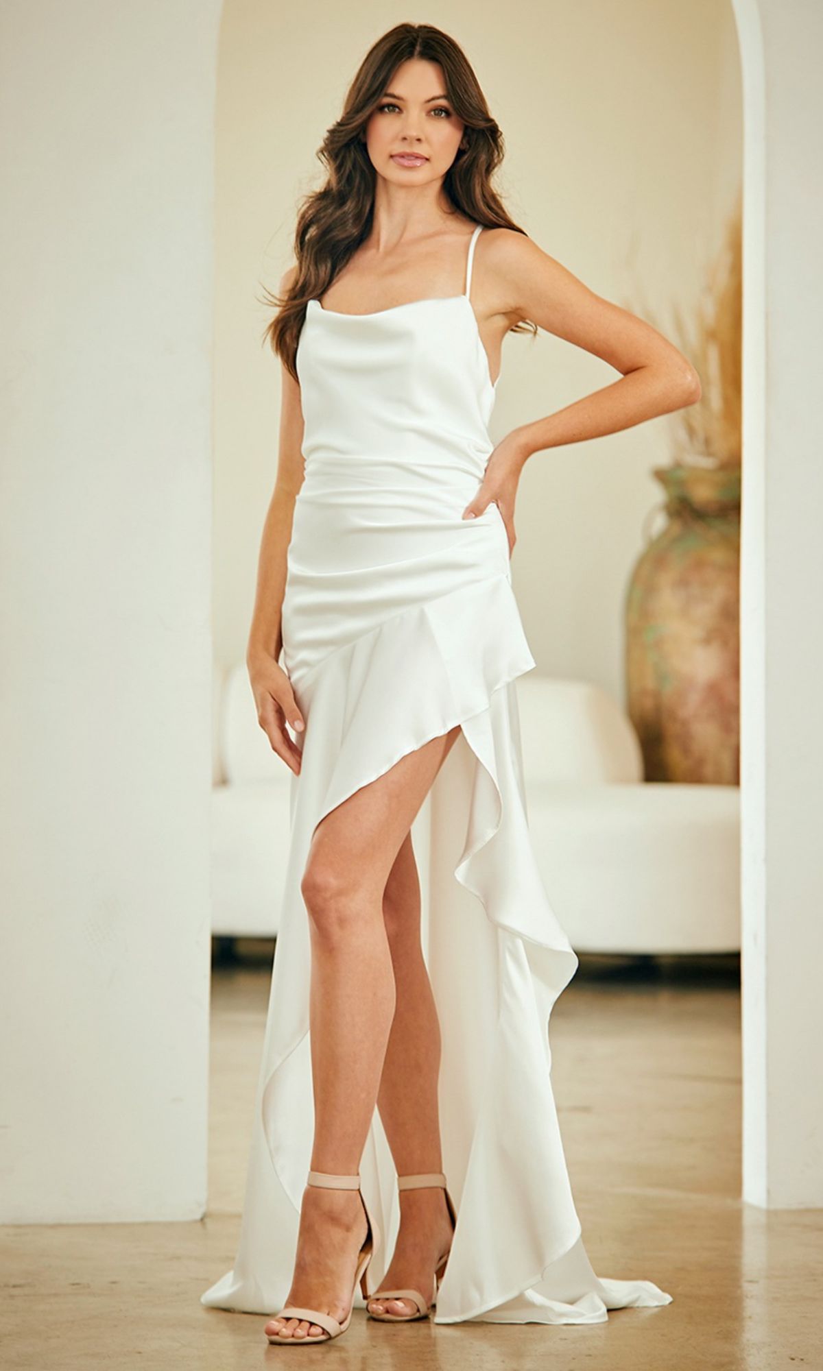 A woman stands indoors in a white Strappy-Back Long Prom Dress (Amelia Couture 6131) with an asymmetrical high slit and train, nude heels, and loose wavy hair.