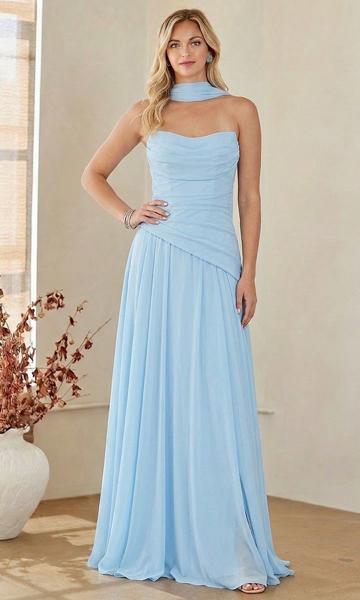 A woman with long blond hair stands indoors wearing the Amelia Couture 6133 strapless chiffon prom gown in light blue, featuring a ruched corset and matching neck scarf, accessorized with bracelets.