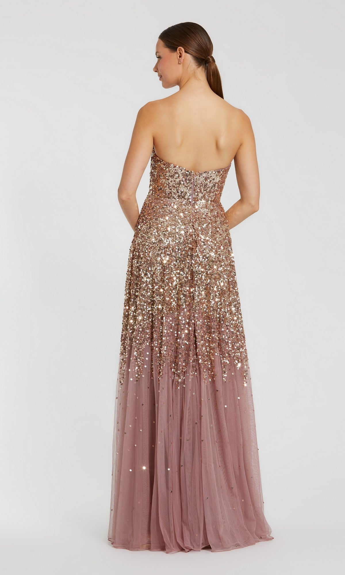 A woman stands with her back to the camera, modeling the Mac Duggal 6144 long formal dress—a strapless gown adorned with rose gold ombre sequins on the bodice and a sheer, floor-length tulle skirt.