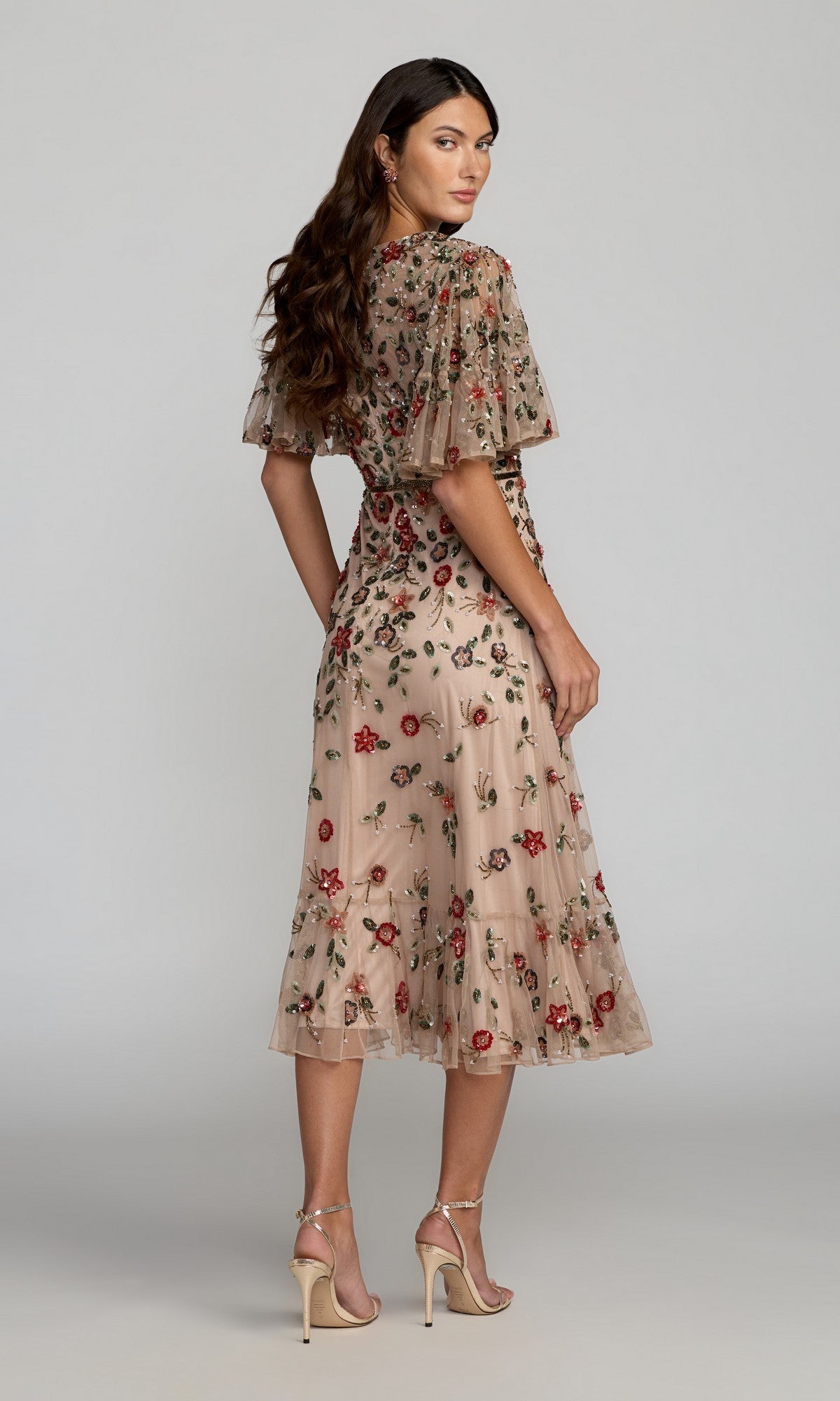 Woman in the Mac Duggal 6162 Green Midi Wedding Guest Dress with red and green floral embroidery, short sheer sleeves, and nude high-heeled sandals, standing and looking over her shoulder.