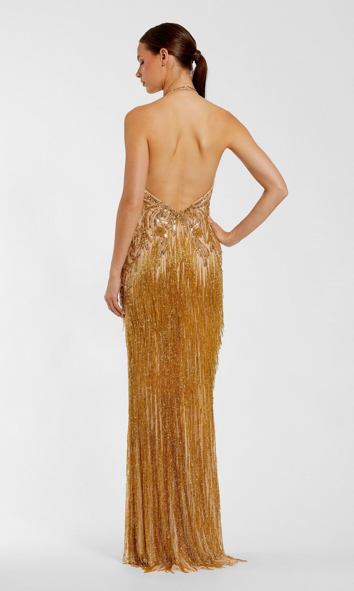 A woman stands facing away against a plain white background, her hair styled in a low ponytail, wearing the Mac Duggal 6170 Long Formal Dress—a gold, sequin-embellished, backless evening gown.