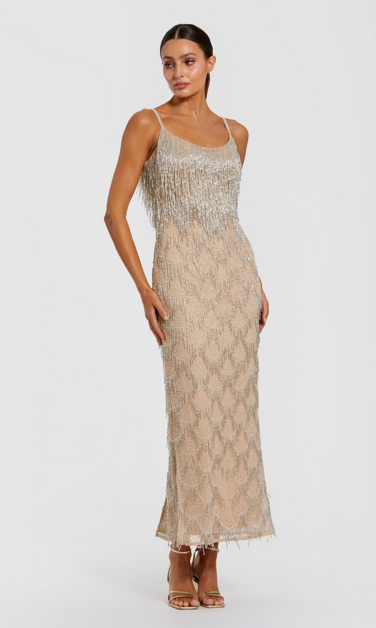 A woman wearing the Mac Duggal 6172 long formal dress in Nude Silver, a sleeveless midi design adorned with shimmering fringe and sequin details, stands in heels against a plain white background.