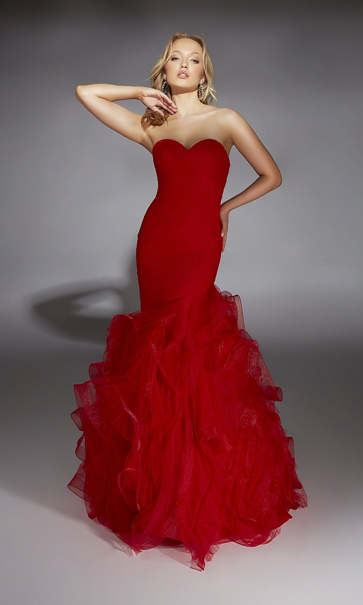 A person poses in the Alyce 61739 long prom dress, a strapless mermaid gown with a sweetheart neckline and ruffled tulle skirt, its bold red color standing out against a neutral background.