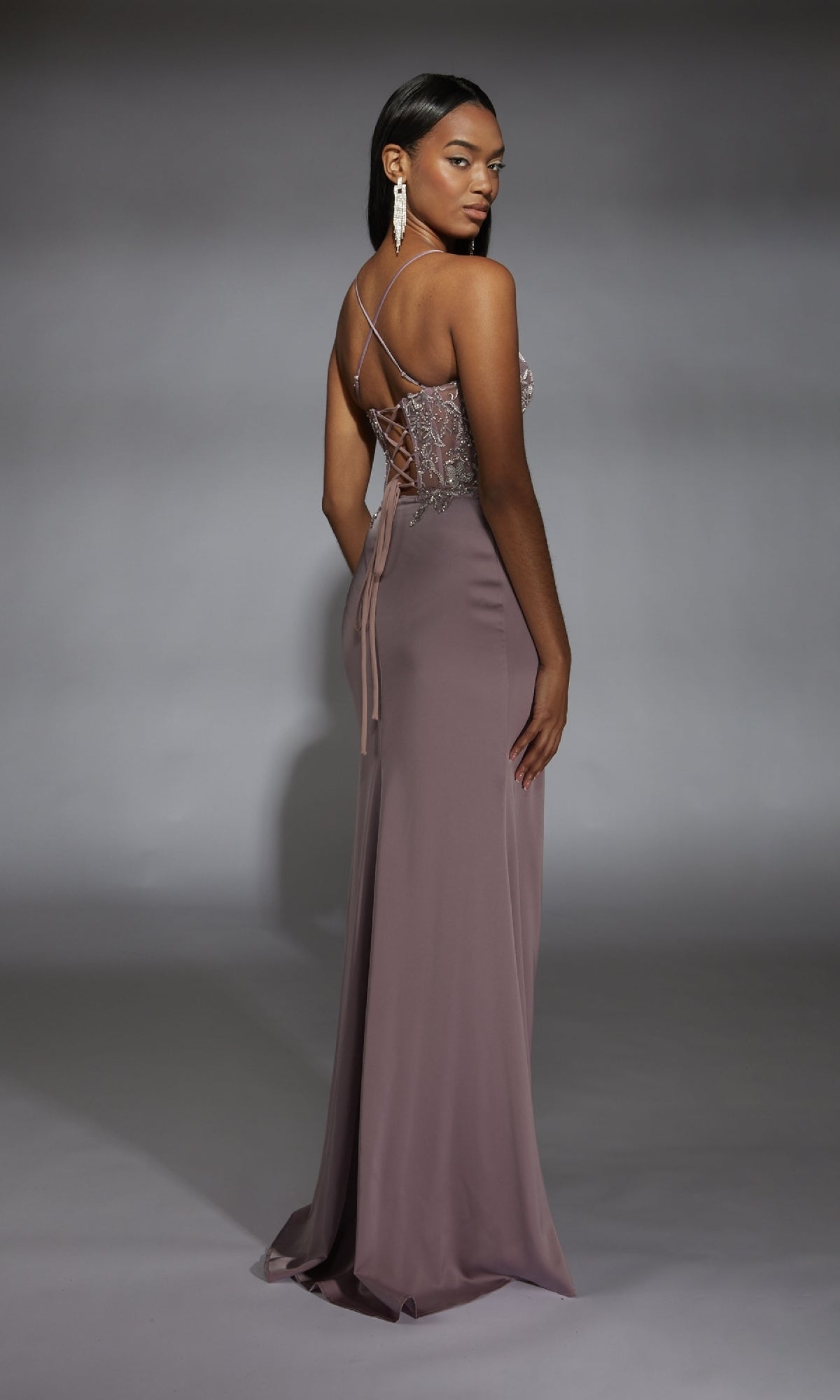 A woman wears the Alyce 61741 Beaded-Lace-Corset Long Prom Dress with a beaded bodice, lace-up back, and thin straps, standing in a studio setting and facing away from the camera.