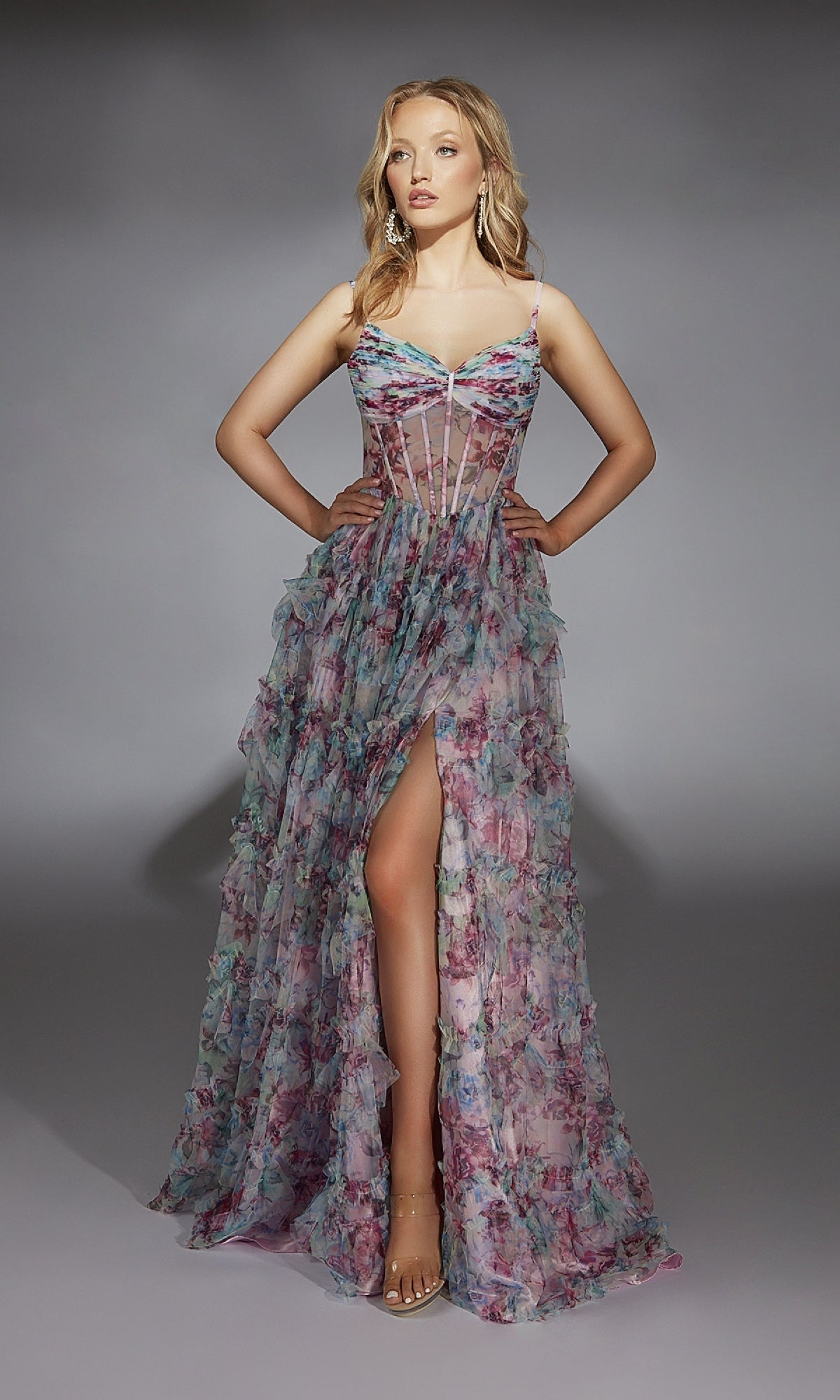 A woman stands in a studio against a plain gray background, wearing the Alyce 61748 long prom dress—a sleeveless, sheer floral-patterned gown with a corset bodice, high-leg slit, and spaghetti straps.