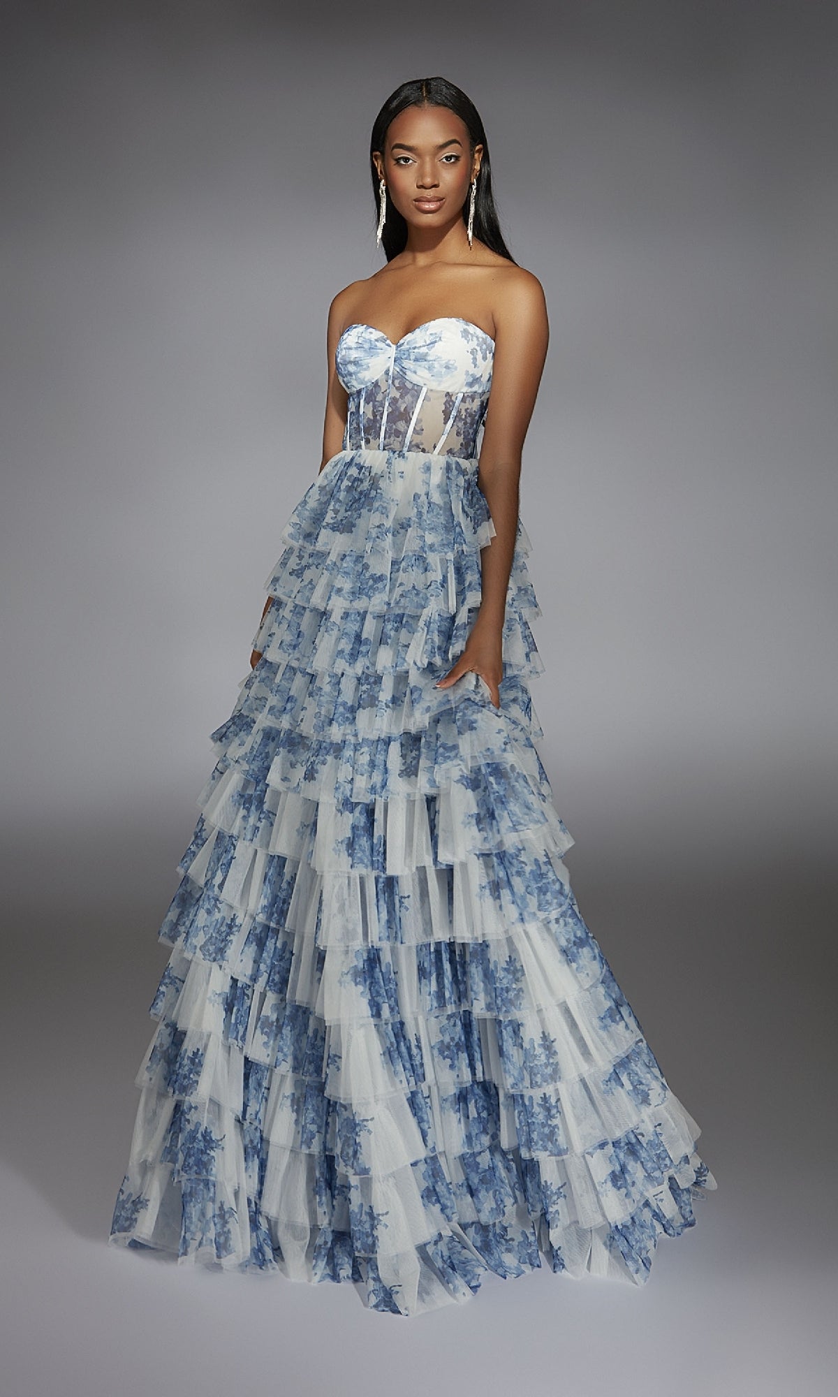 A woman stands wearing the Alyce 61763 long prom dress, which features a strapless sheer corset bodice and a layered ruffled skirt in blue floral print tulle.