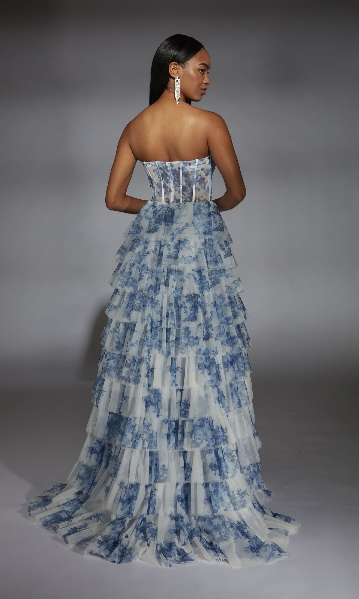 Model wears the Alyce 61763 long prom dress, a strapless design with a blue floral print and sheer corset bodice, styled with long earrings and photographed from the back against a gray backdrop.