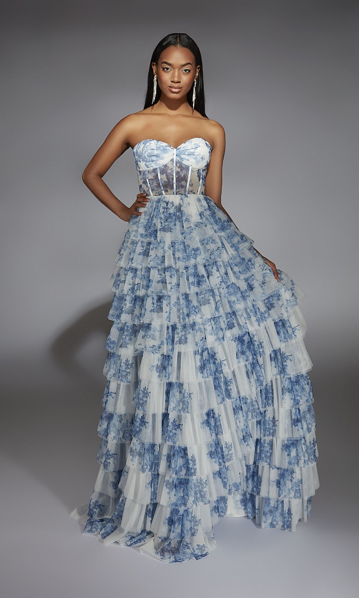 A woman wears the Alyce 61763 long prom dress, featuring a strapless sheer corset bodice and tiered ruffled skirt, all in a blue floral print.