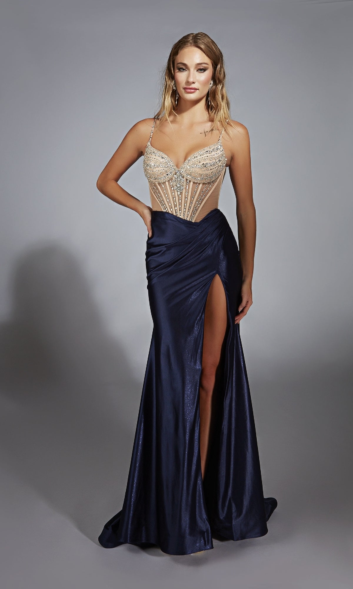 A woman stands against a plain gray background wearing the Alyce 61767 long navy blue prom dress made of stretch satin, featuring a sheer beaded bodice and a high leg slit.