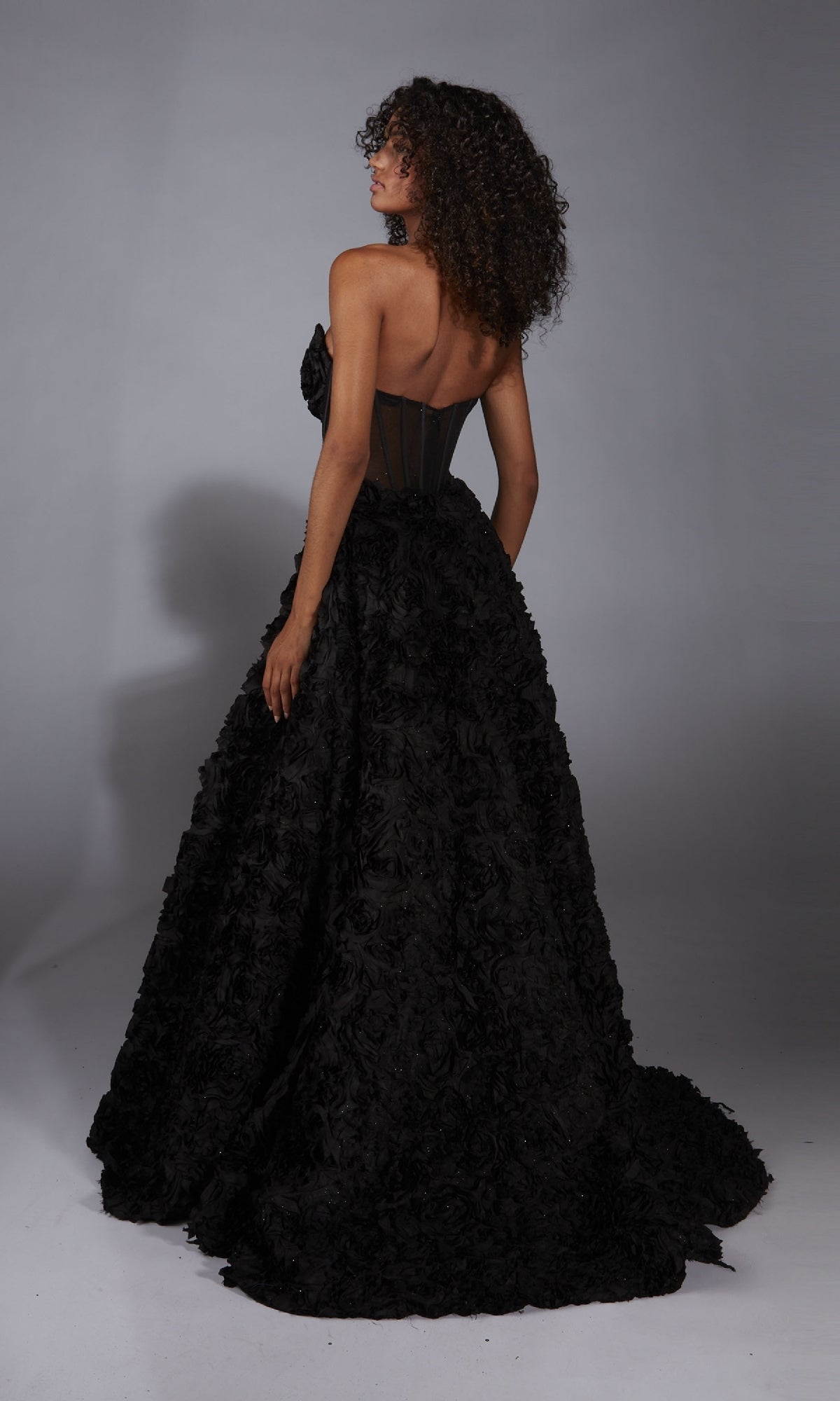 A person with curly hair wears the Alyce 61775 Strapless Rosette Prom Ball Gown, featuring a corset-style bodice and textured, voluminous skirt, standing facing away in a studio.