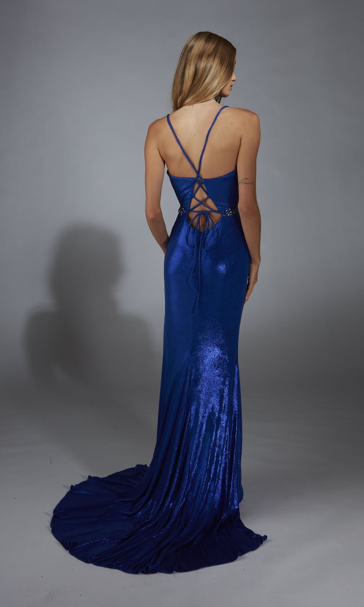 A woman faces away, wearing the Alyce 61785 long blue prom dress with a shiny metallic finish, lace-up back, and slight train, set against a plain gray background.