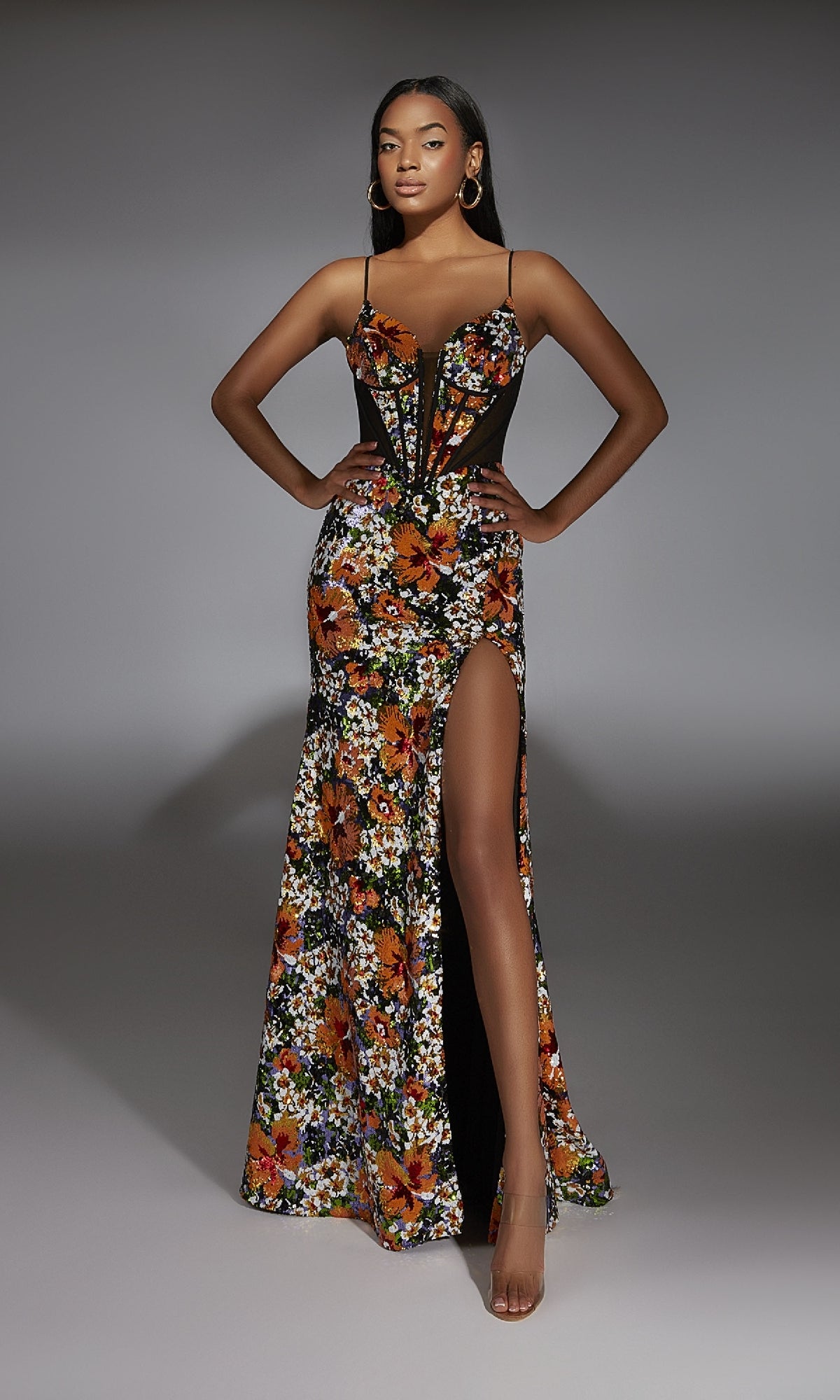 A woman poses with hands on hips in the Alyce 61809 long prom dress—a sleeveless floral gown with a corset bodice, high slit, and black mesh accents—against a plain gray background.