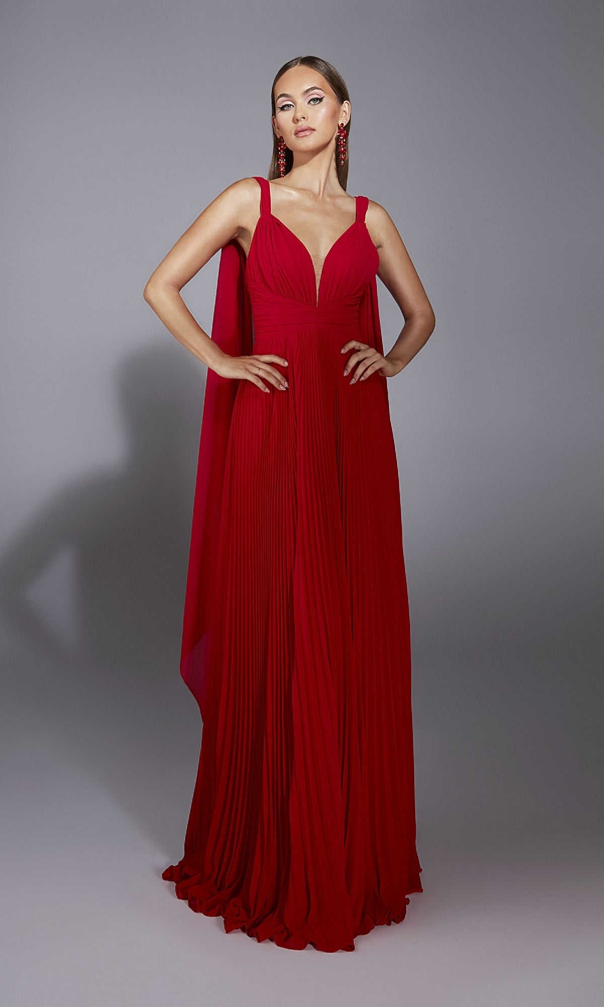 A woman stands in a studio against a gray background wearing the Alyce 61823 long red prom dress with pleats, deep V-neckline, and matching red earrings, her hands on her hips.