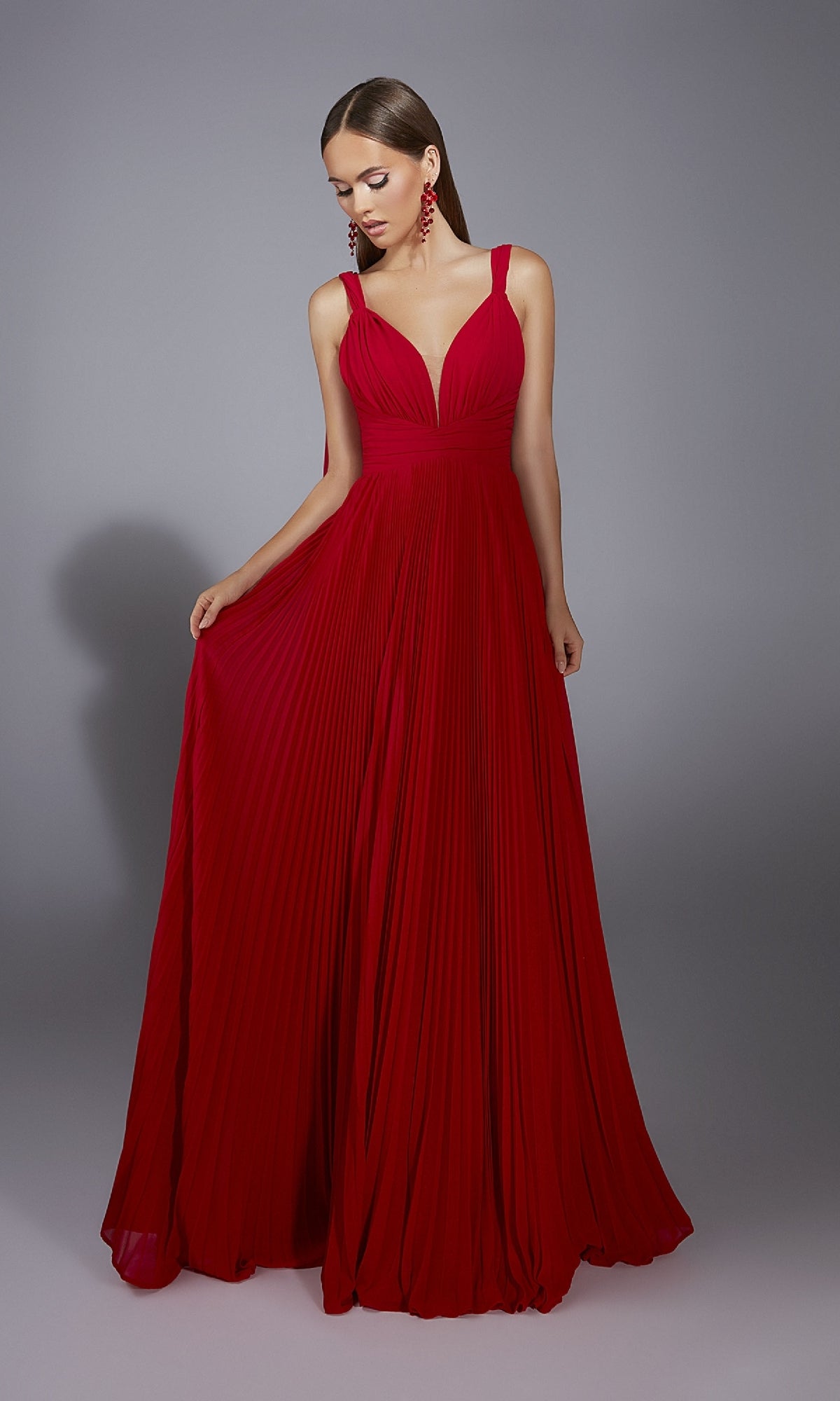 Model wearing the Alyce 61823 long prom dress—floor-length, sleeveless, with a deep v-neckline and pleated A-line skirt—in red, standing against a plain gray background.