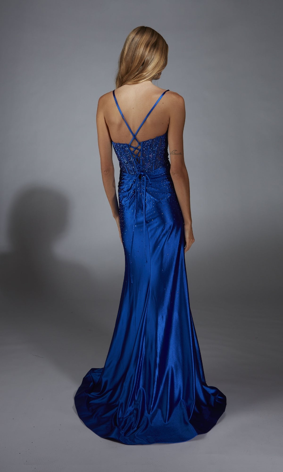A woman in the Alyce 61827 Beaded-Corset Long Prom Dress, featuring a royal blue satin finish, thin crisscross straps, and a floor-length sleeveless design, stands facing away from the camera.