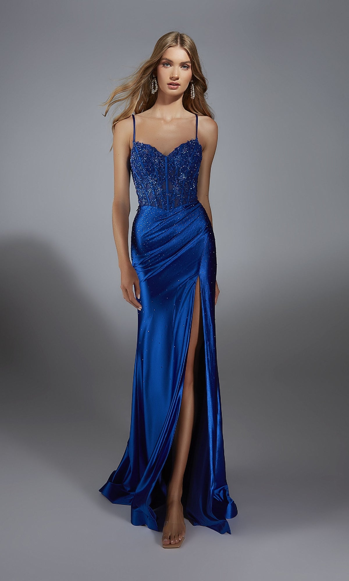 A woman in the Alyce 61827 Beaded-Corset Long Prom Dress—a blue, floor-length gown with a beaded bodice, thin straps, and a high slit—stands against a plain gray background.