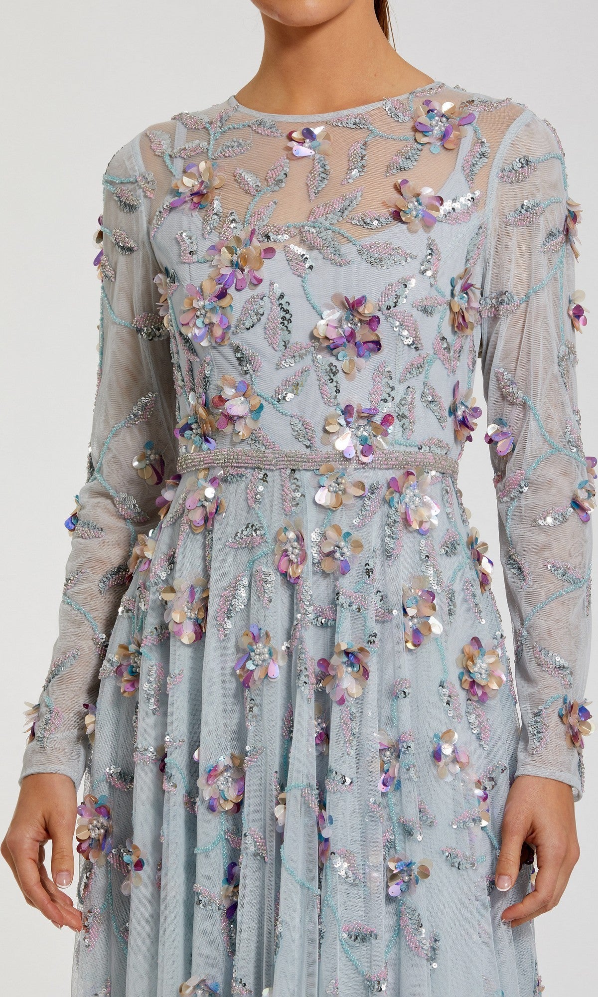 A woman wearing the Mac Duggal 6183 Long Sleeve Beaded Formal Gown—an elegant light blue sheer dress featuring embroidered floral patterns and 3D sequin flower embellishments.